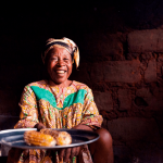 closeup of old african lady in her rural home kitchen with traditional dress looking in camera offering food